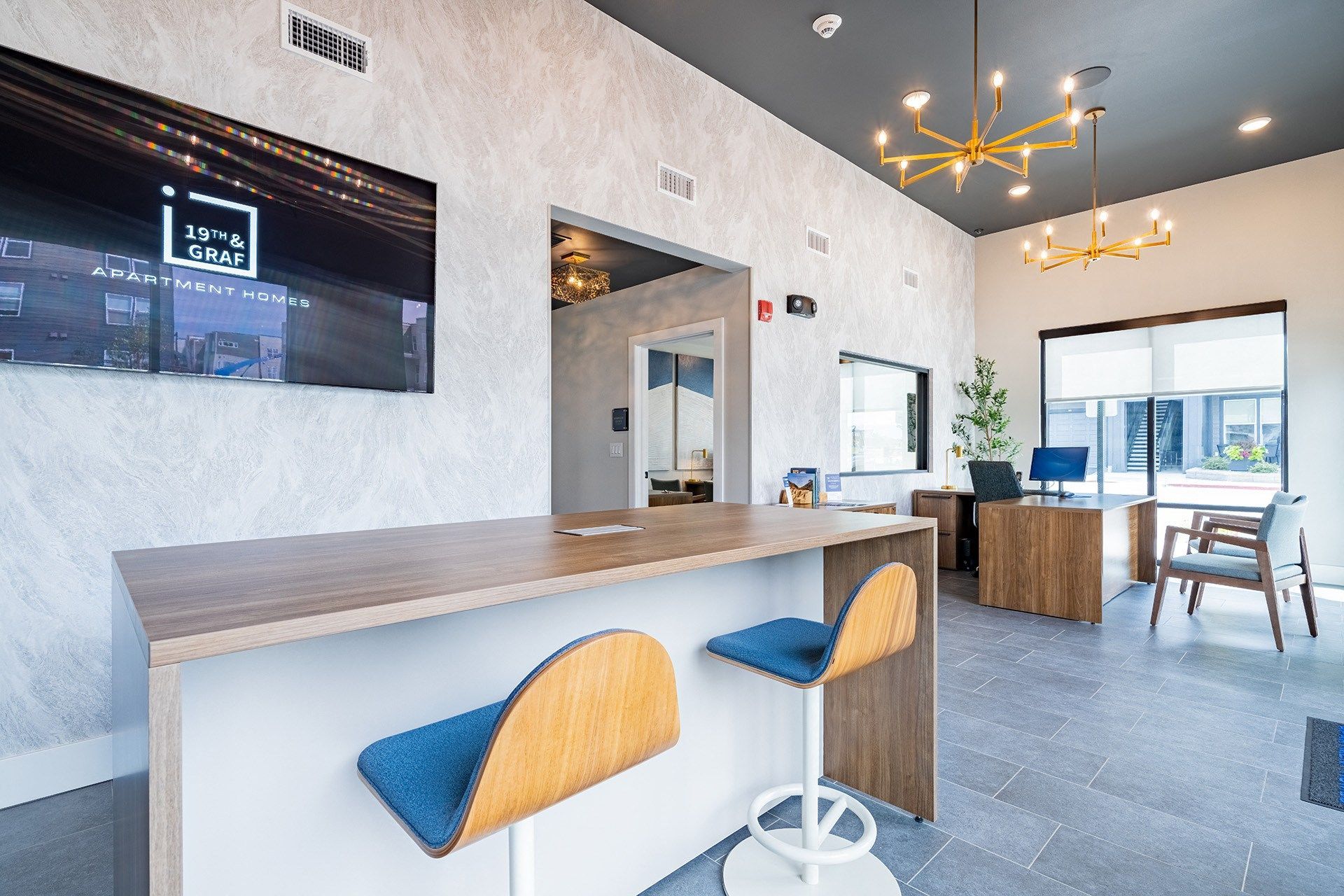 19th and Graf  Apartments Lobby with a bar, stools, and a desk. TV on the wall. Light fixtures hang from the ceiling.