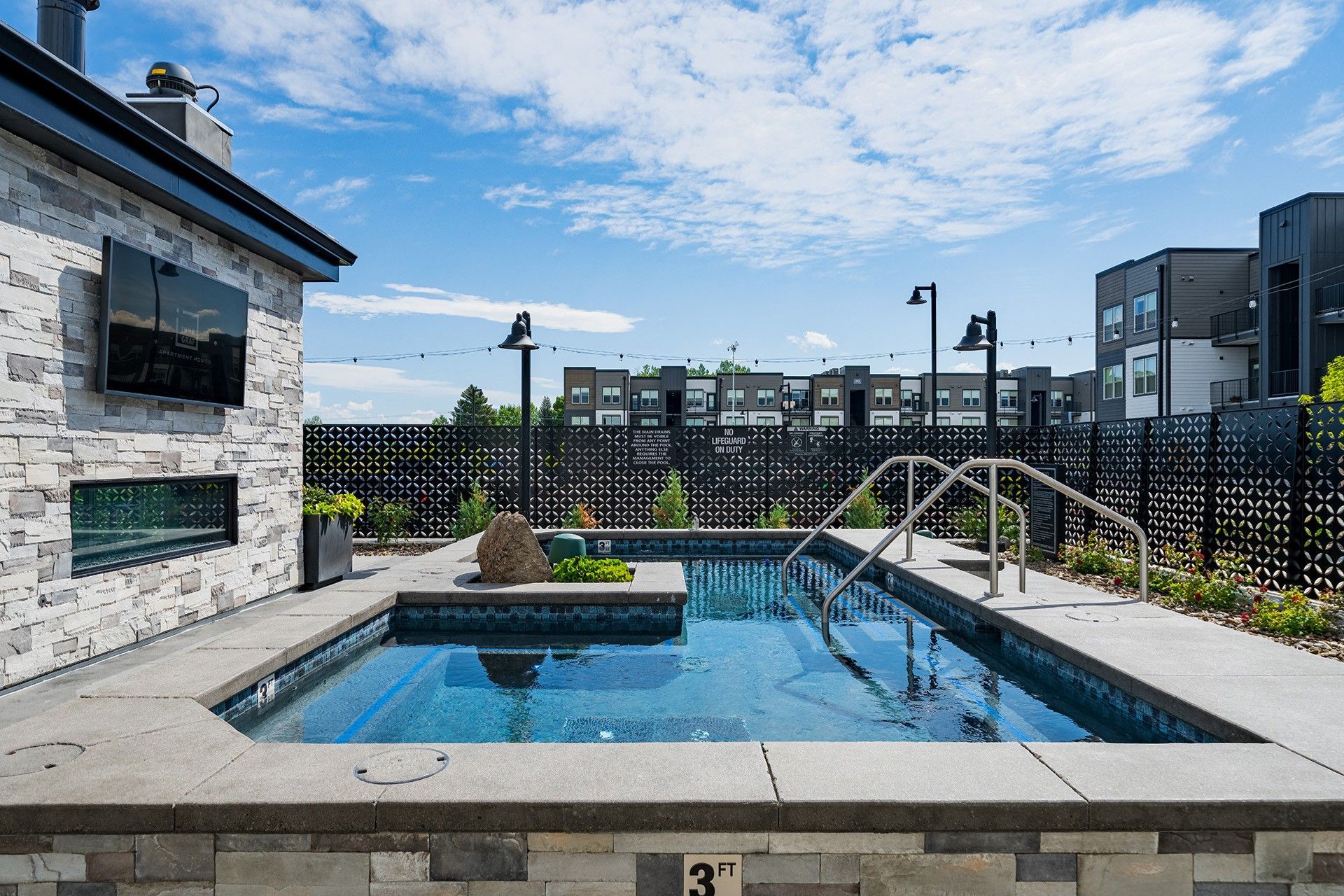 19th and Graf  Apartments Small outdoor pool with stone surround, metal fence, and apartment buildings in the background under a blue sky.