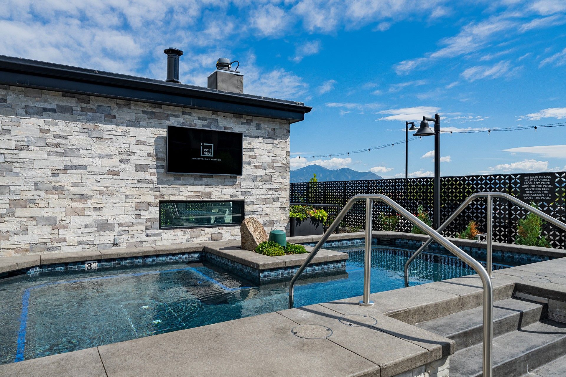 19th and Graf  Apartments Outdoor pool with stone wall, steps, and handrails. TV mounted on the wall. Blue sky with clouds.