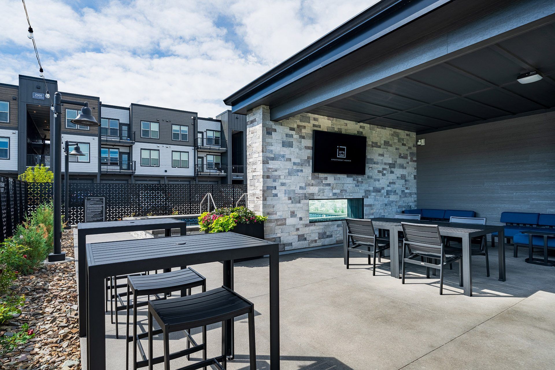 19th and Graf  Apartments Outdoor lounge area with tables, chairs, and a TV mounted on a stone wall. Apartment building in the background.