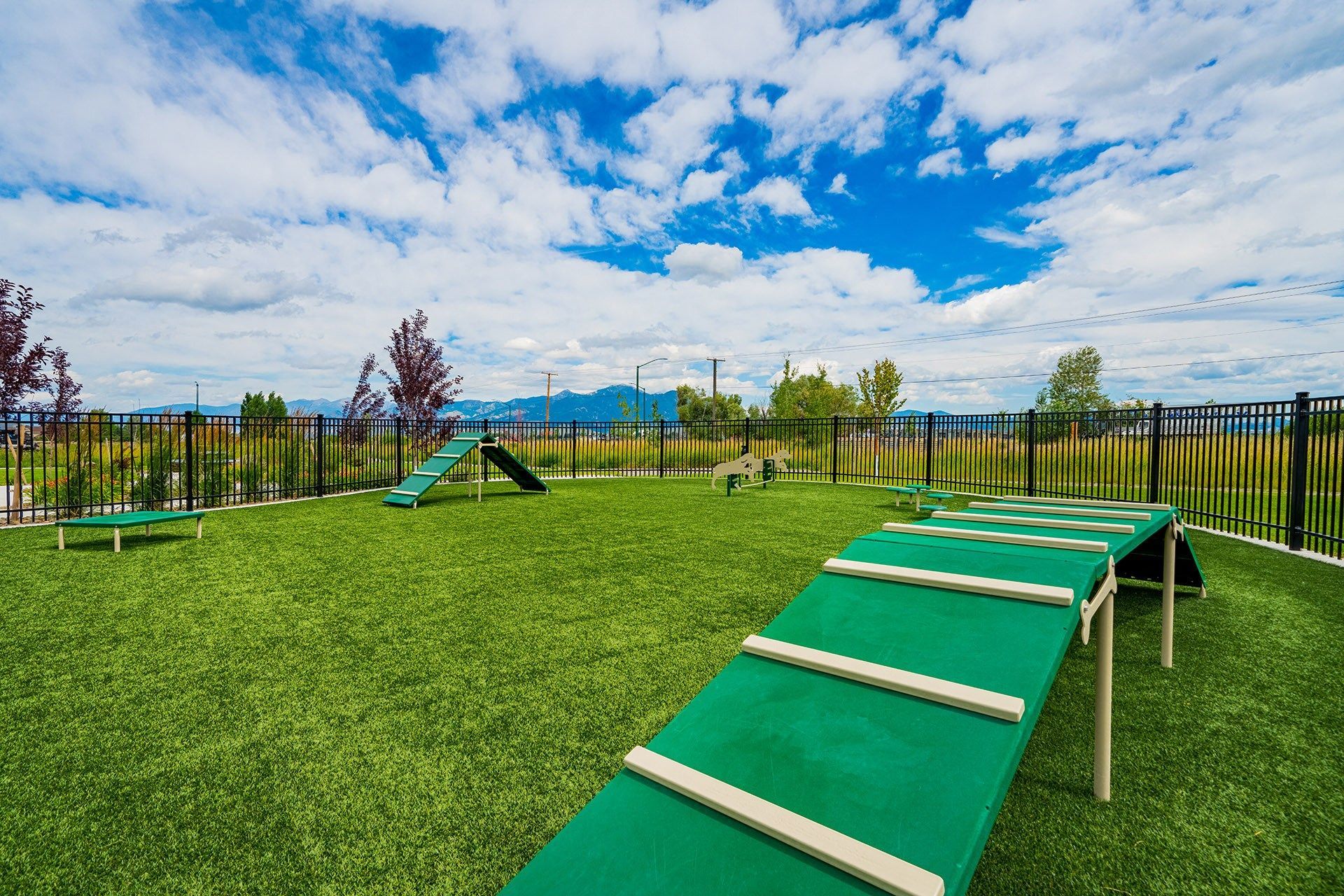 19th and Graf  Apartments Dog park with green artificial turf, agility equipment, black fence, blue sky, and clouds.