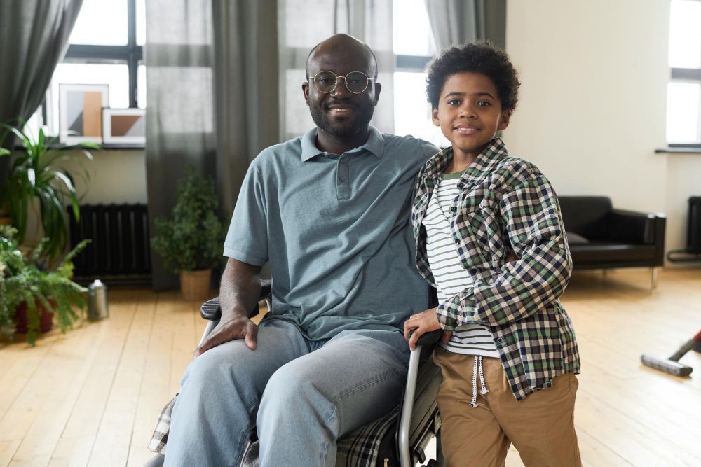 Two people in a bright living room, one seated in a wheelchair, smiling beside each other