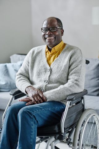 Smiling man seated in a wheelchair, wearing a gray cardigan and yellow shirt in a bright room