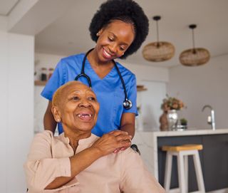 Smiling caregiver in blue scrubs hugging an older woman in a bright home kitchen