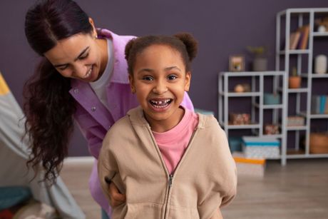 Smiling child in beige hoodie with adult behind her in a playroom