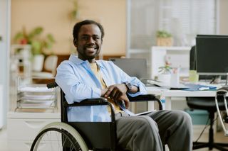Man in a wheelchair smiling in a bright office, sitting at a desk with papers and a computer.