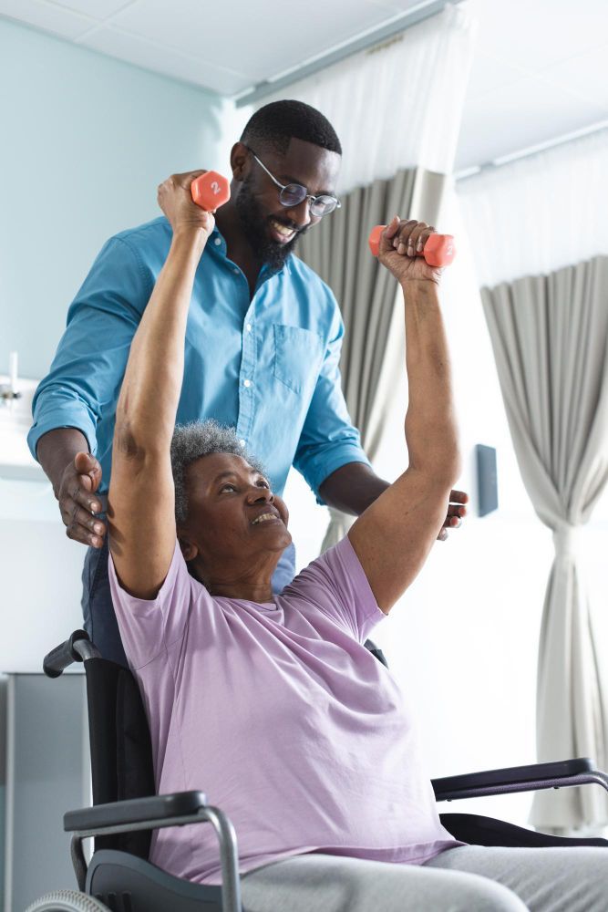Caregiver helping a seated patient lift red dumbbells during physical therapy in a bright room