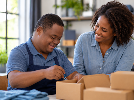 Two people packing cardboard boxes together at a table, smiling in a bright room