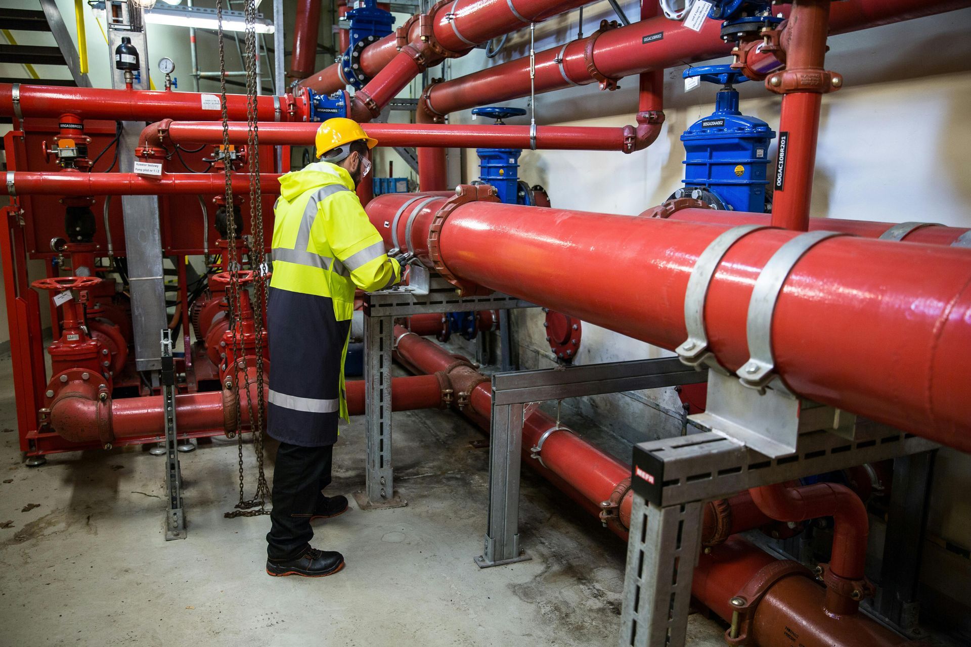 A technician in a high-visibility yellow jacket inspects a complex network of large red industrial pipes indoors.