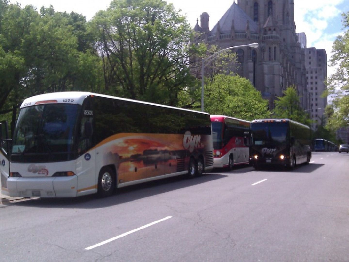 Three tour buses lined up along a tree-lined city street beside a stone church