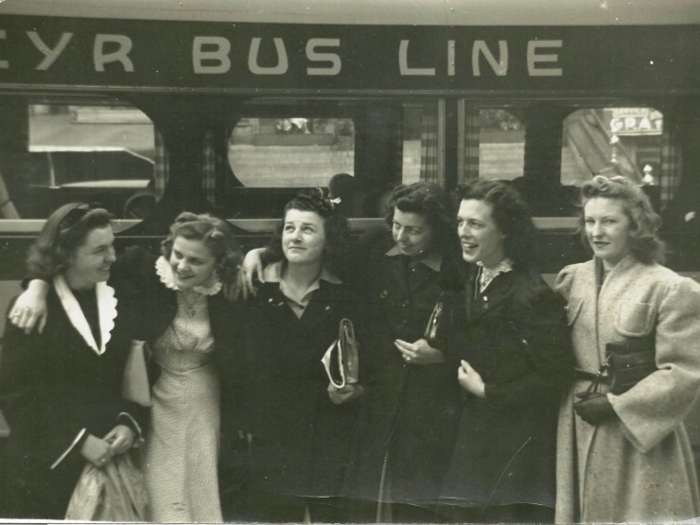 Five people standing outside a bus marked “... BUS LINE,” posing and smiling in coats and dresses.