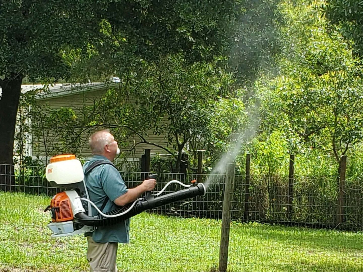 Man spraying a yard with a backpack sprayer. Spray is white against green grass, trees, and a house.