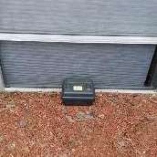 A black utility box sits on the ground in front of a gray garage door. Reddish-brown mulch surrounds it.