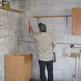 Person spraying a wall in a room. Gray cinder block walls, wooden shelves, and a cabinet are visible.