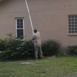 Person using a long-handled tool to work on the exterior of a building; near windows and bushes.