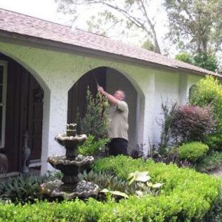 Man spraying water, standing under arched entryway of white stucco building with fountain and bushes in front.