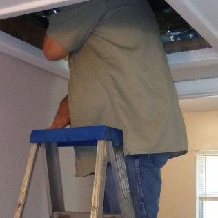 Man on a step ladder, working on ceiling access panel. He's wearing a green shirt and jeans.