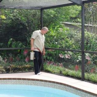 Man spraying insecticide near a pool, surrounded by green foliage, within a screened enclosure.