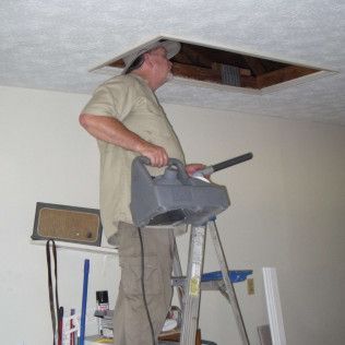 Man on ladder vacuums ceiling opening. Beige shirt, khaki pants, gray vacuum, white ceiling.