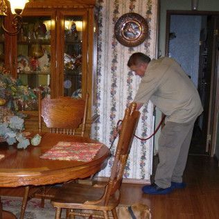 Man vacuuming a room with wooden furniture and patterned wallpaper; he wears blue shoe covers.