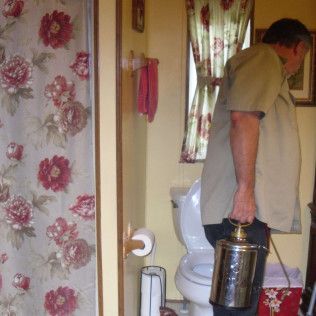Man in bathroom carrying a large metal container; shower curtain with floral pattern.