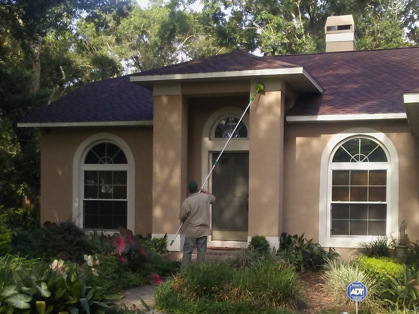 Person using a pole to clean the exterior of a tan house with a red roof, surrounded by plants.