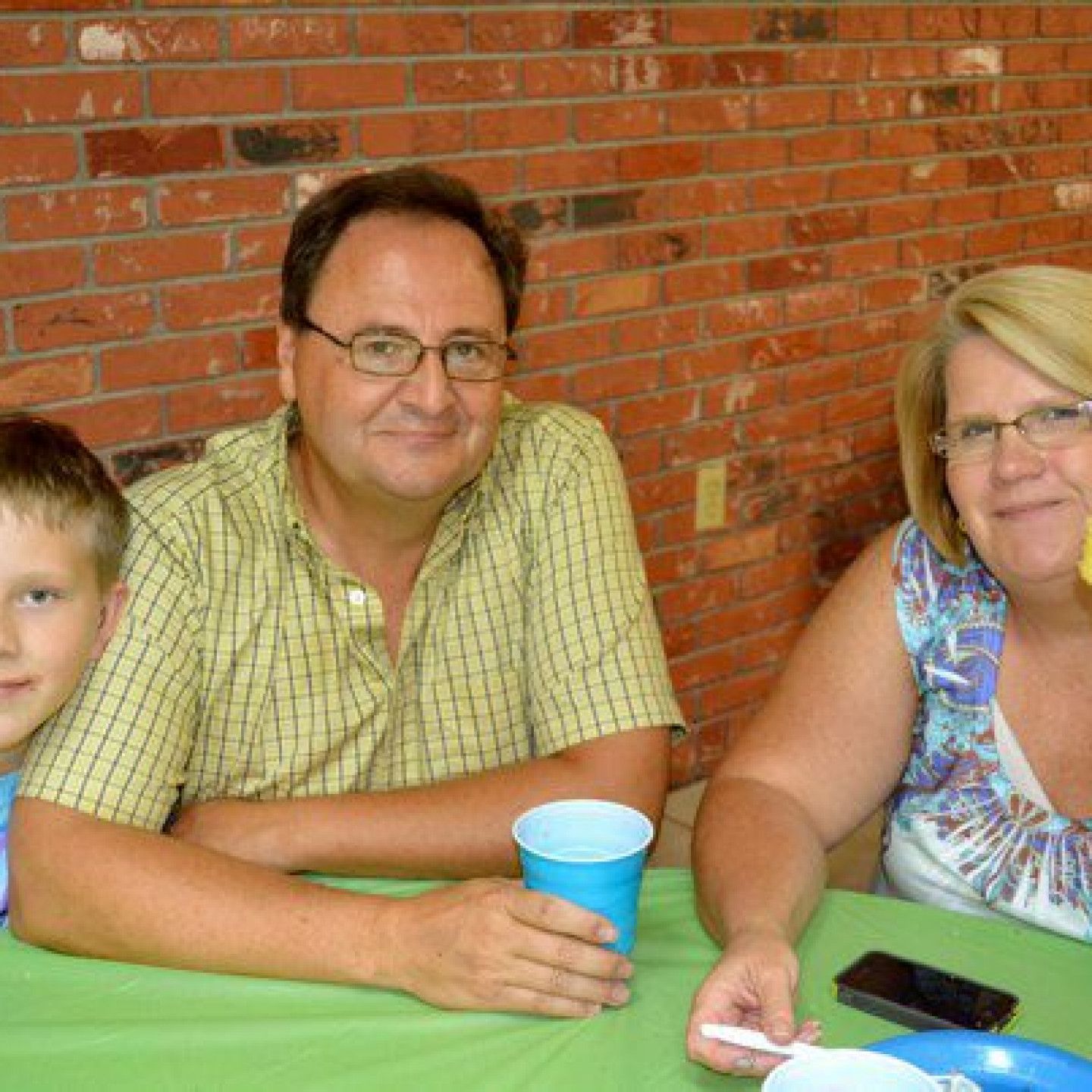 Man and woman seated at a table with a child; brick wall background. Man holds a blue cup.