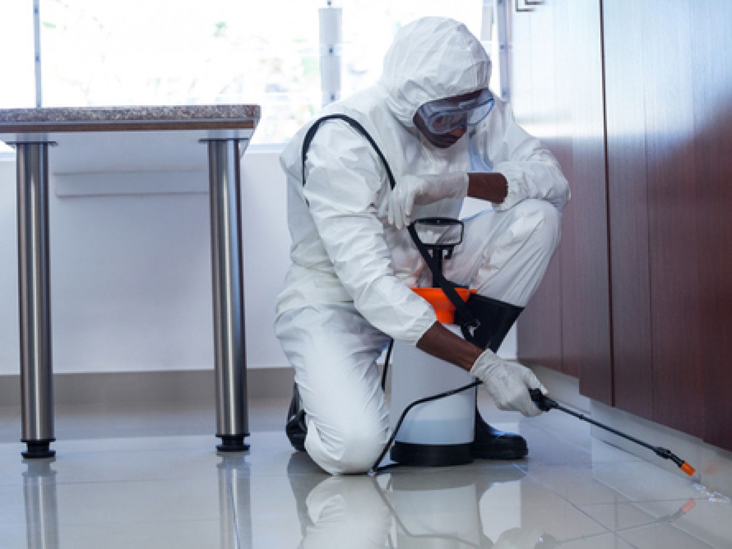 Pest control worker in protective suit kneels, spraying baseboard in a kitchen.