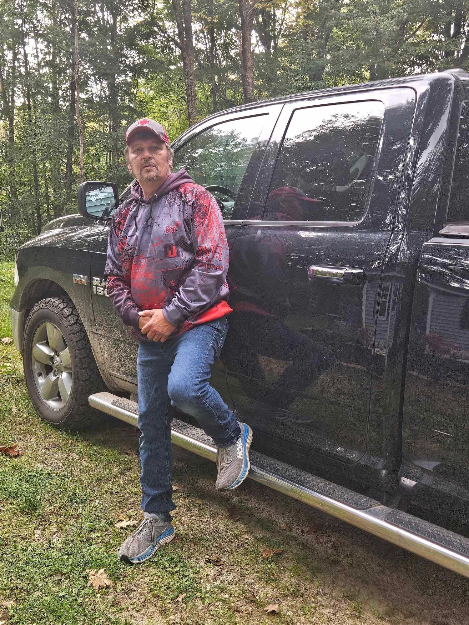 Man in red jacket leaning against a black pickup truck on a wooded dirt path