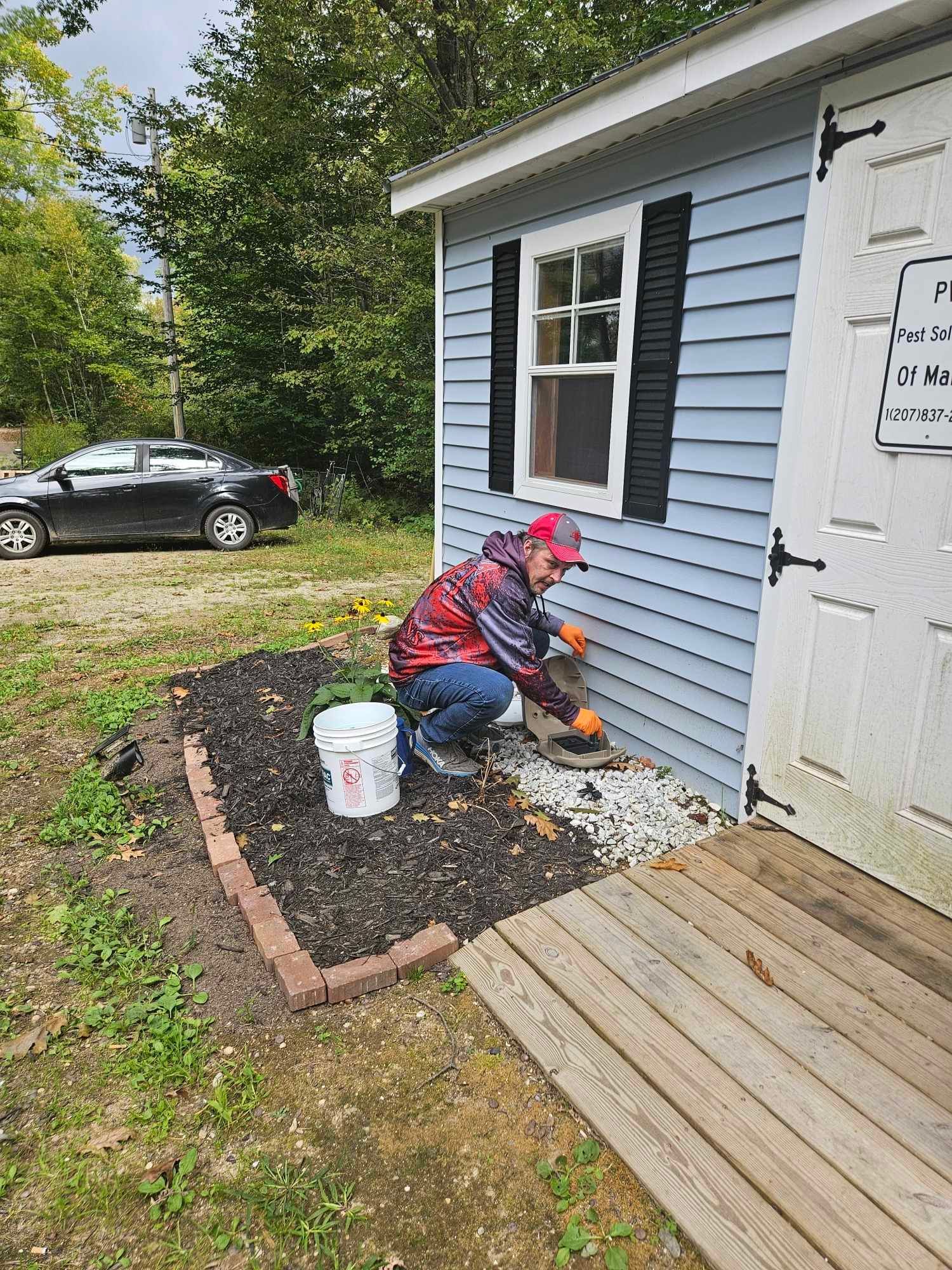 Person in pink jacket laying stones beside a blue house, with a bucket and small walkway.