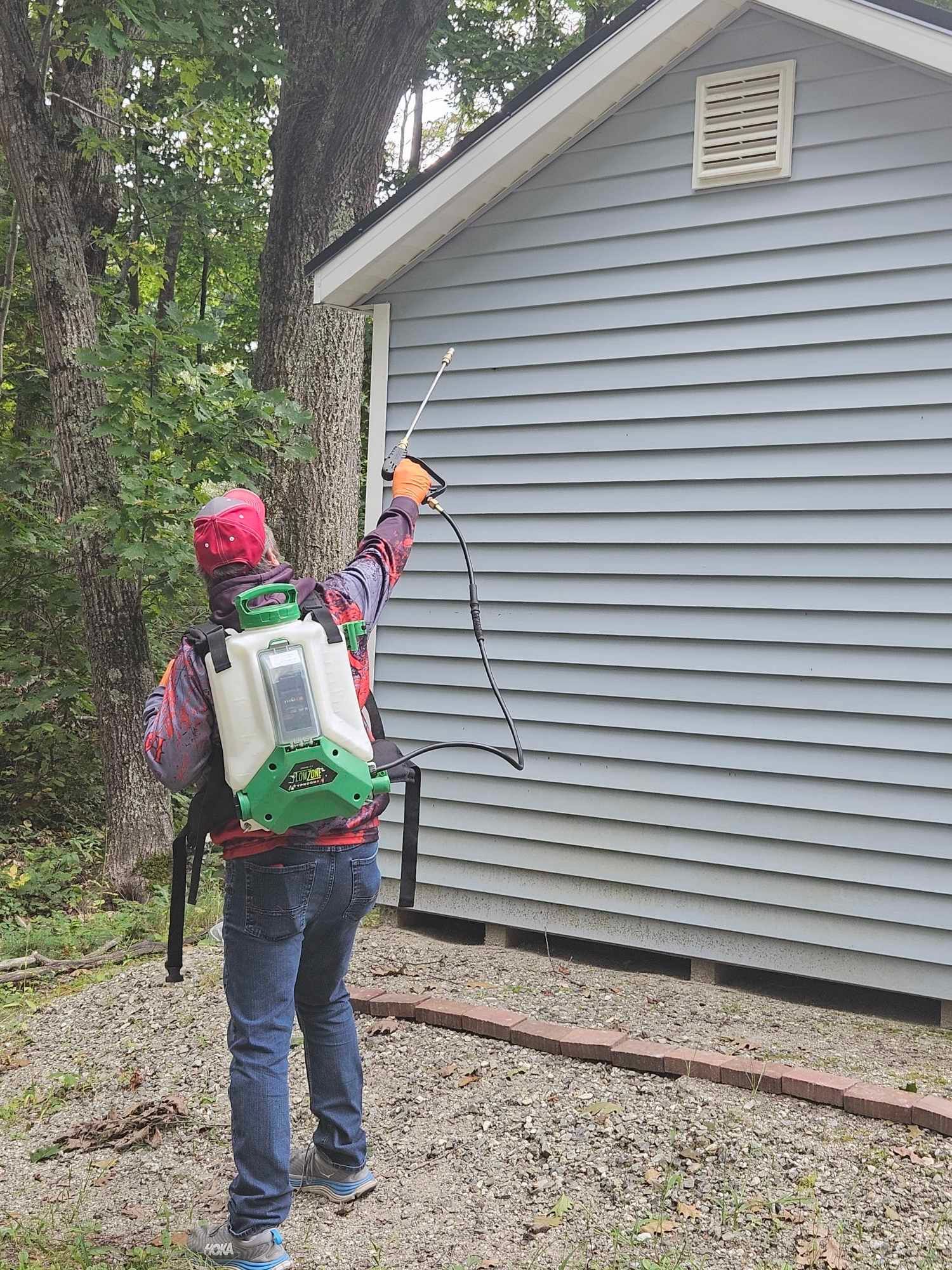 Person with backpack sprayer treating gray house siding outdoors near trees