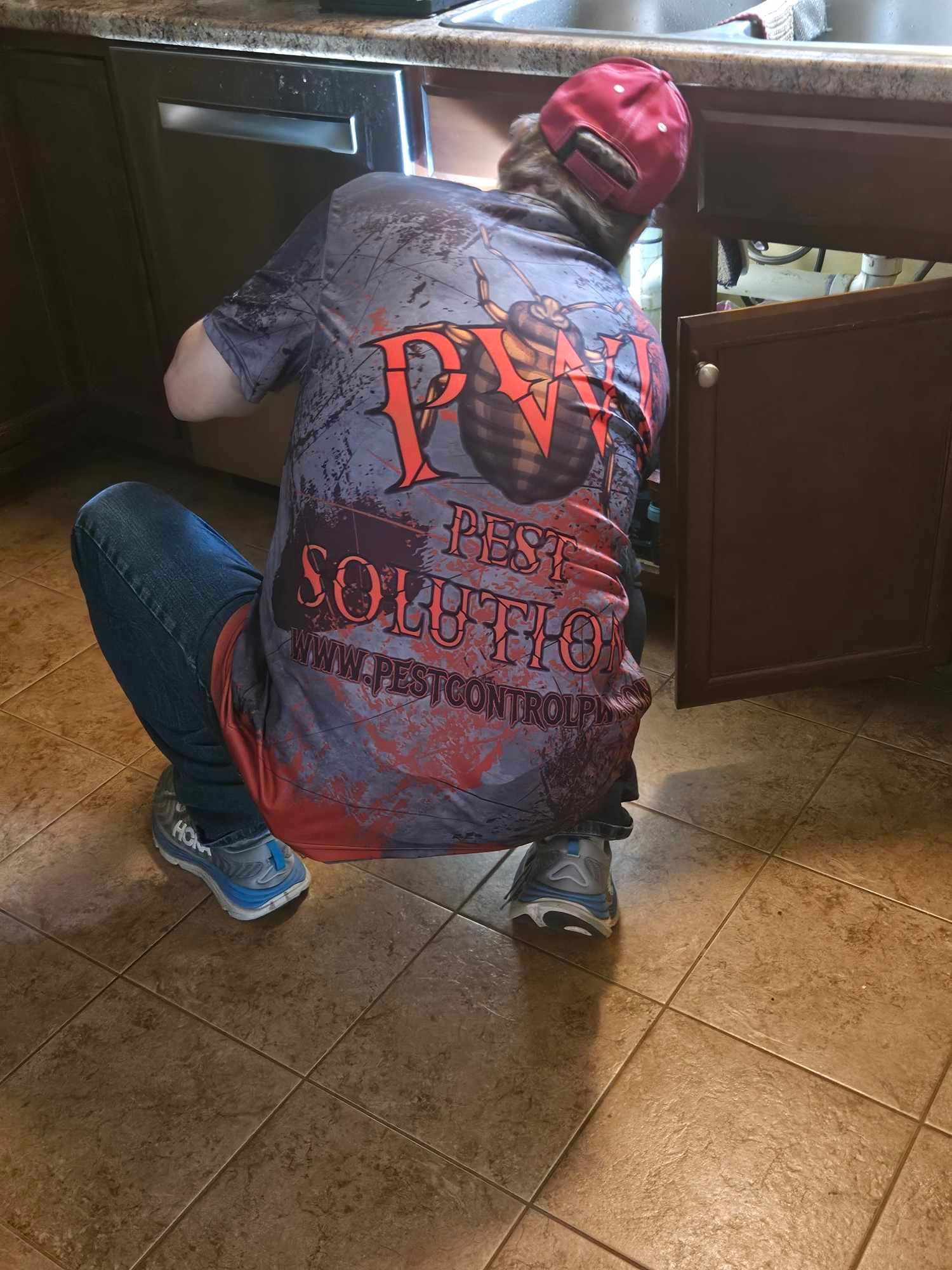 Person crouching by a kitchen cabinet, reaching toward the floor, wearing a red cap and gray shirt with red text.