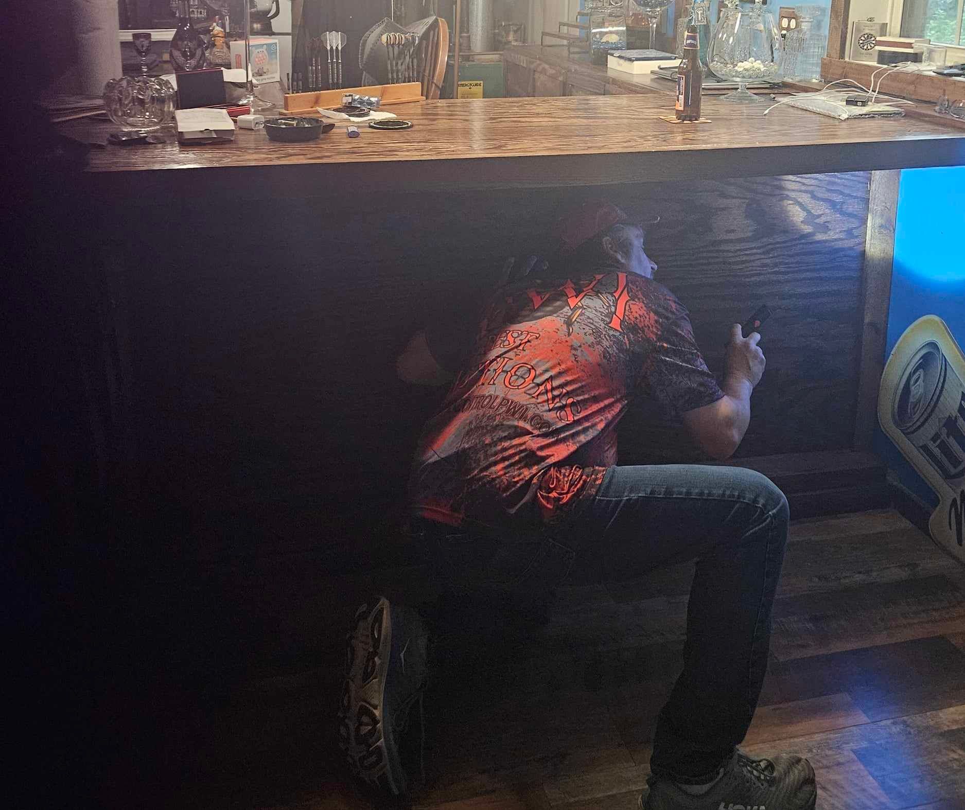 Person crouches under a wooden bar counter, using a phone in a dimly lit room.