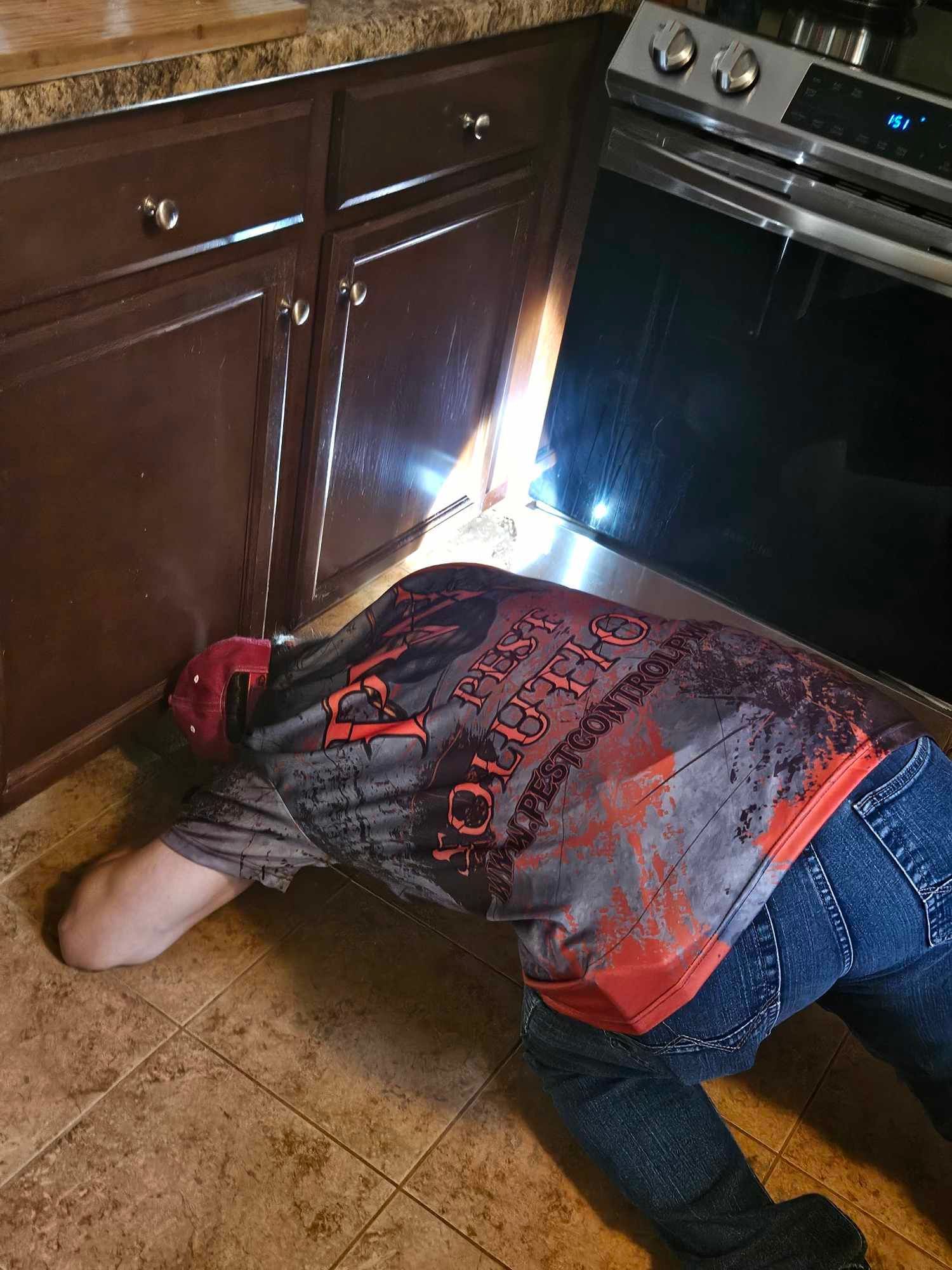Person crouched on a kitchen floor beside cabinets, reaching under a stove with a flashlight.