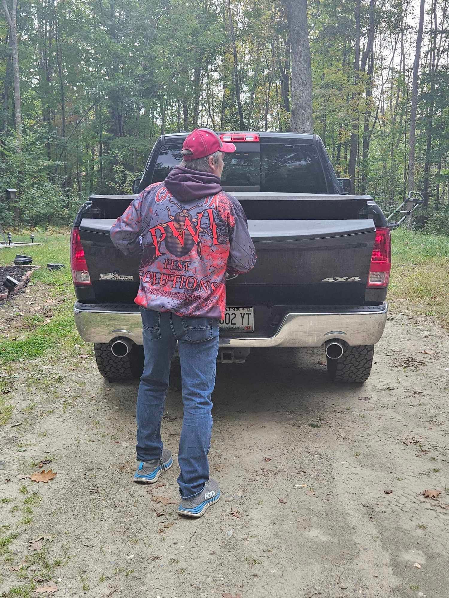 Person standing behind a black pickup truck on a dirt road in a wooded area.