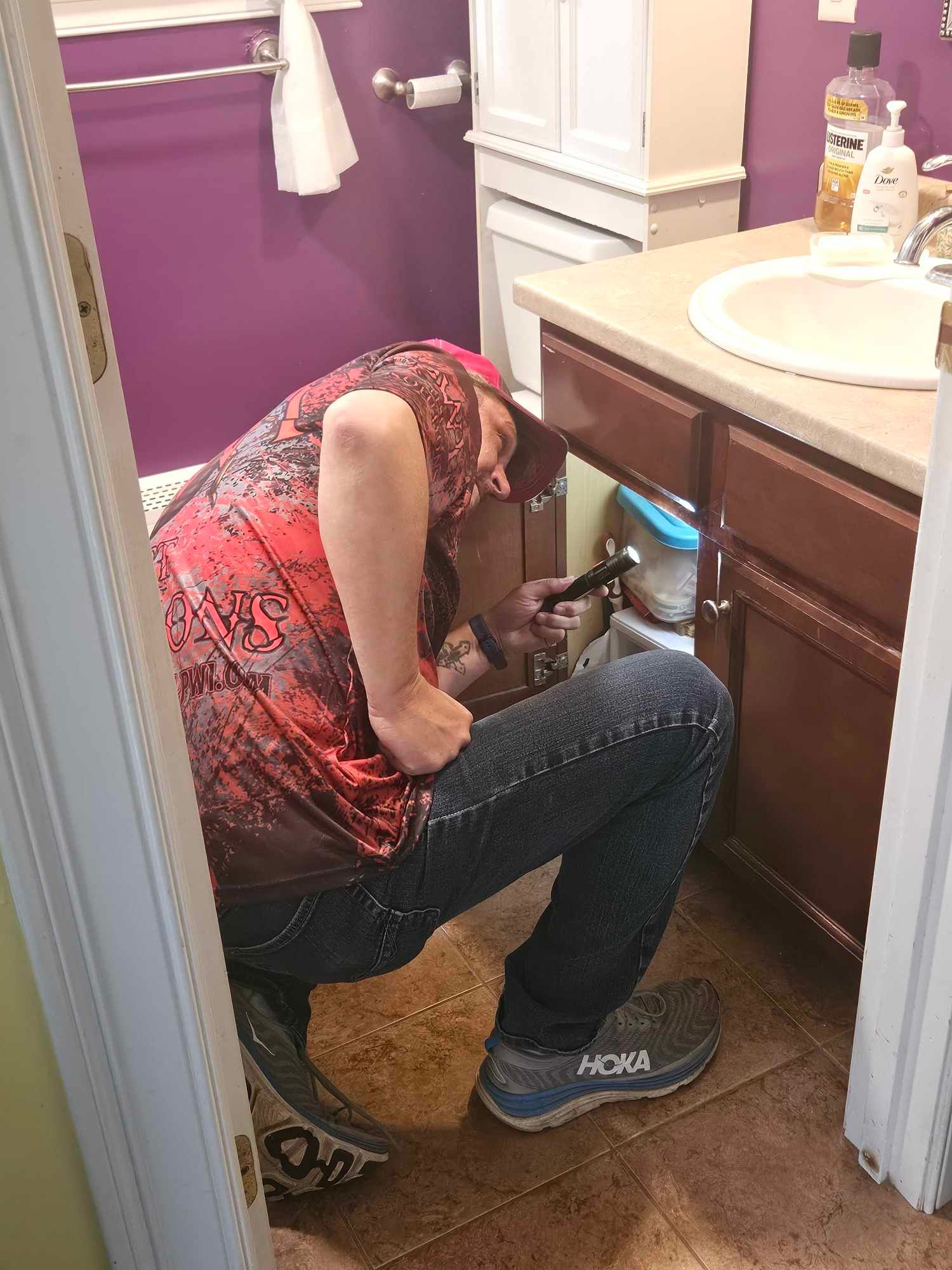 Person crouches under a bathroom sink, working on plumbing in a purple-walled bathroom.