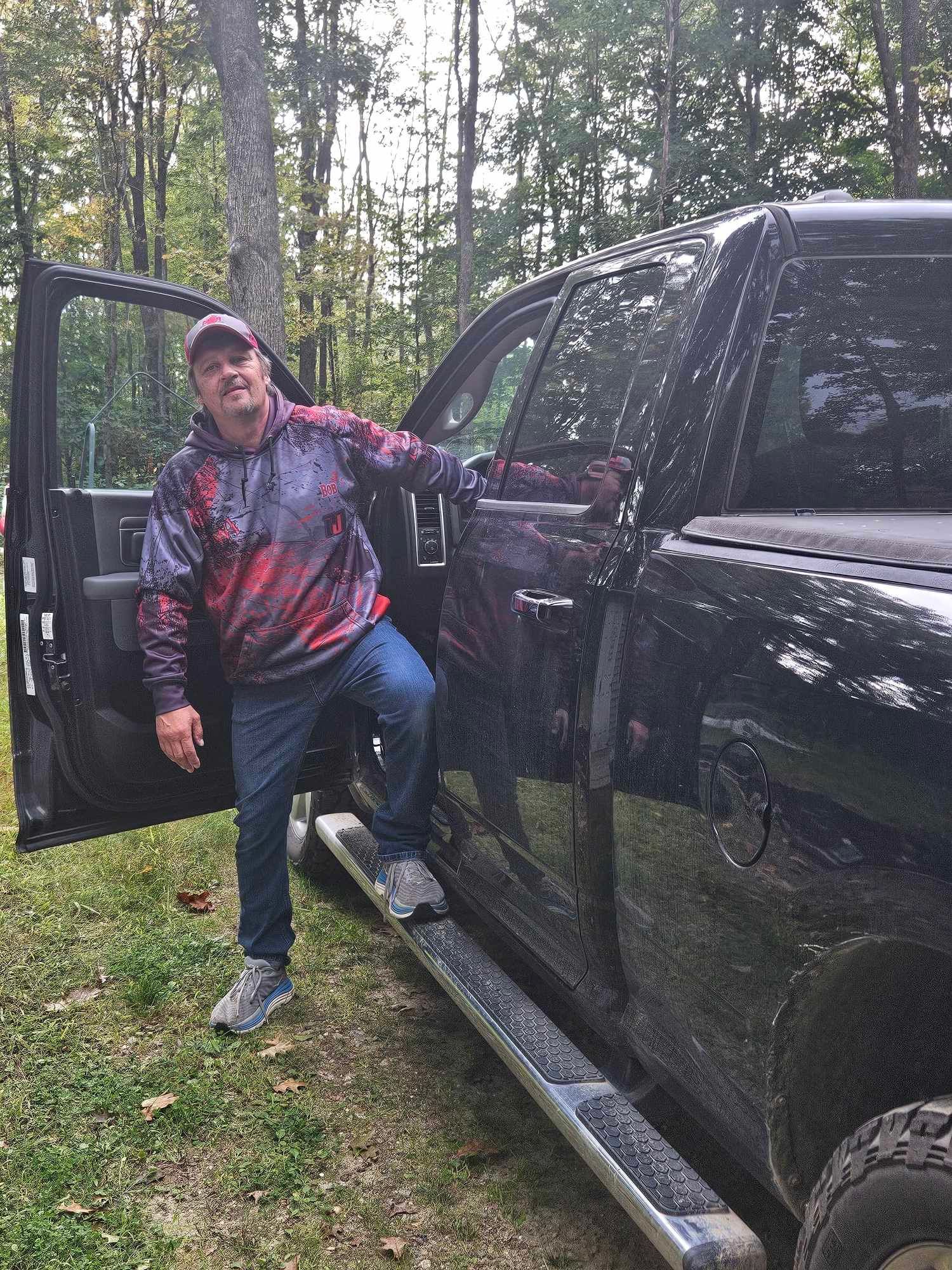 Person standing by an open black pickup truck in a wooded area, wearing a red-and-blue hoodie.