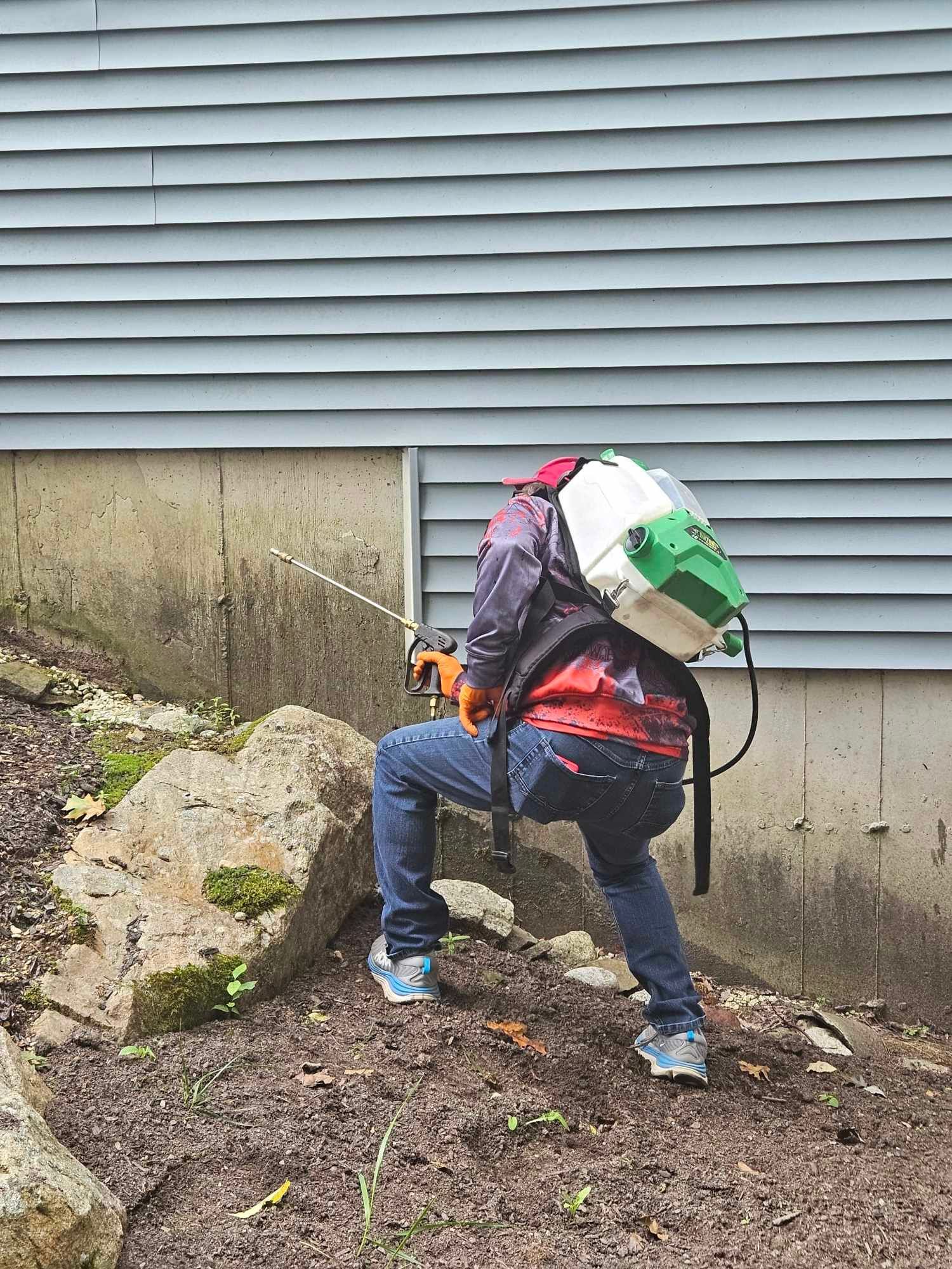 Person carrying a large backpack climbs a muddy slope beside a house with gray siding.