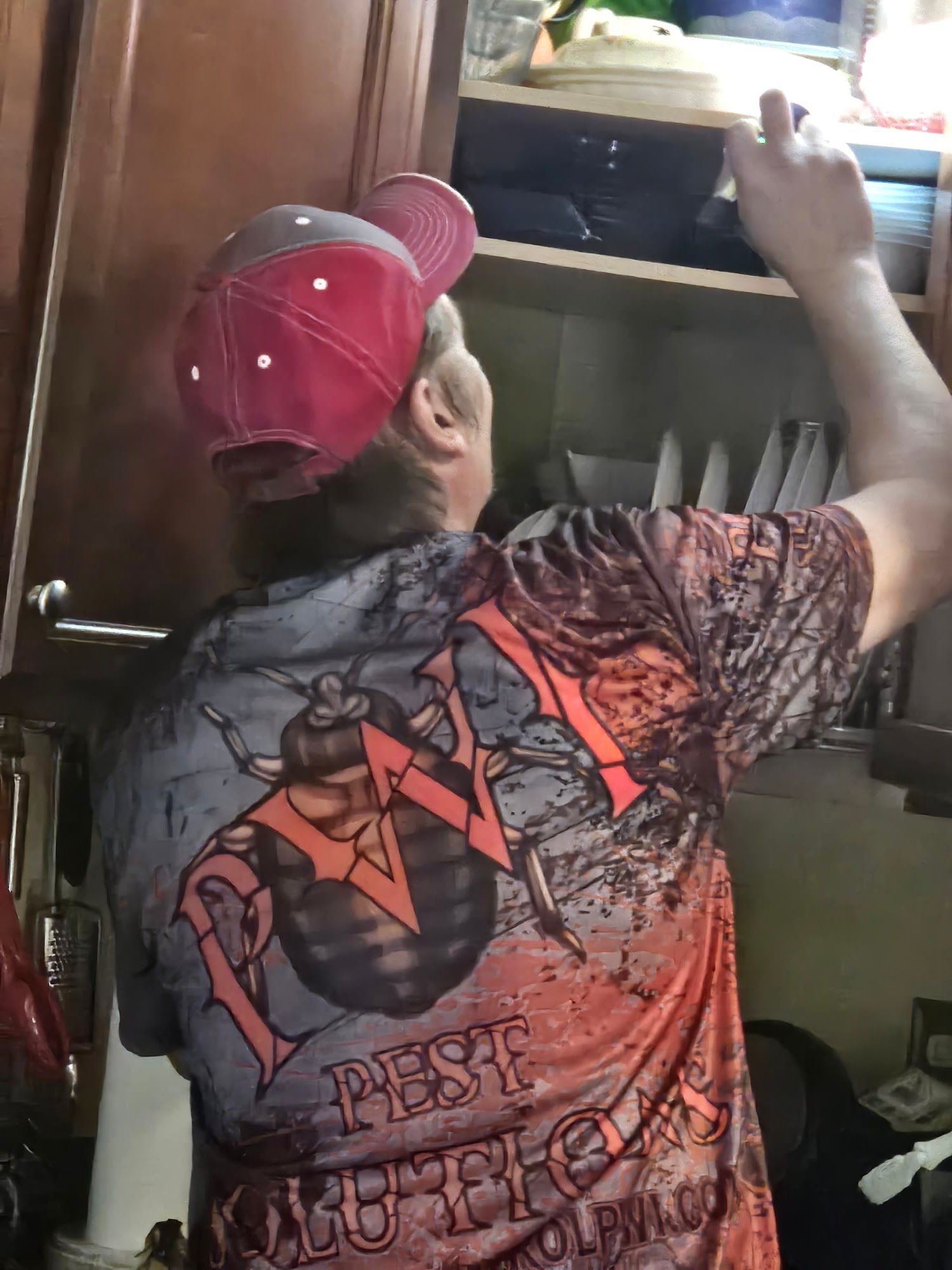 Person in red cap reaching into a kitchen cabinet, wearing a patterned black shirt with red lettering.