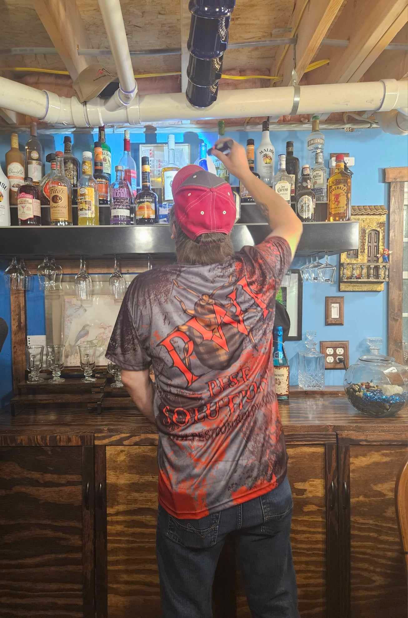 Person in red cap reaches for bottles behind a wooden home bar, with shelves of liquor and glassware.