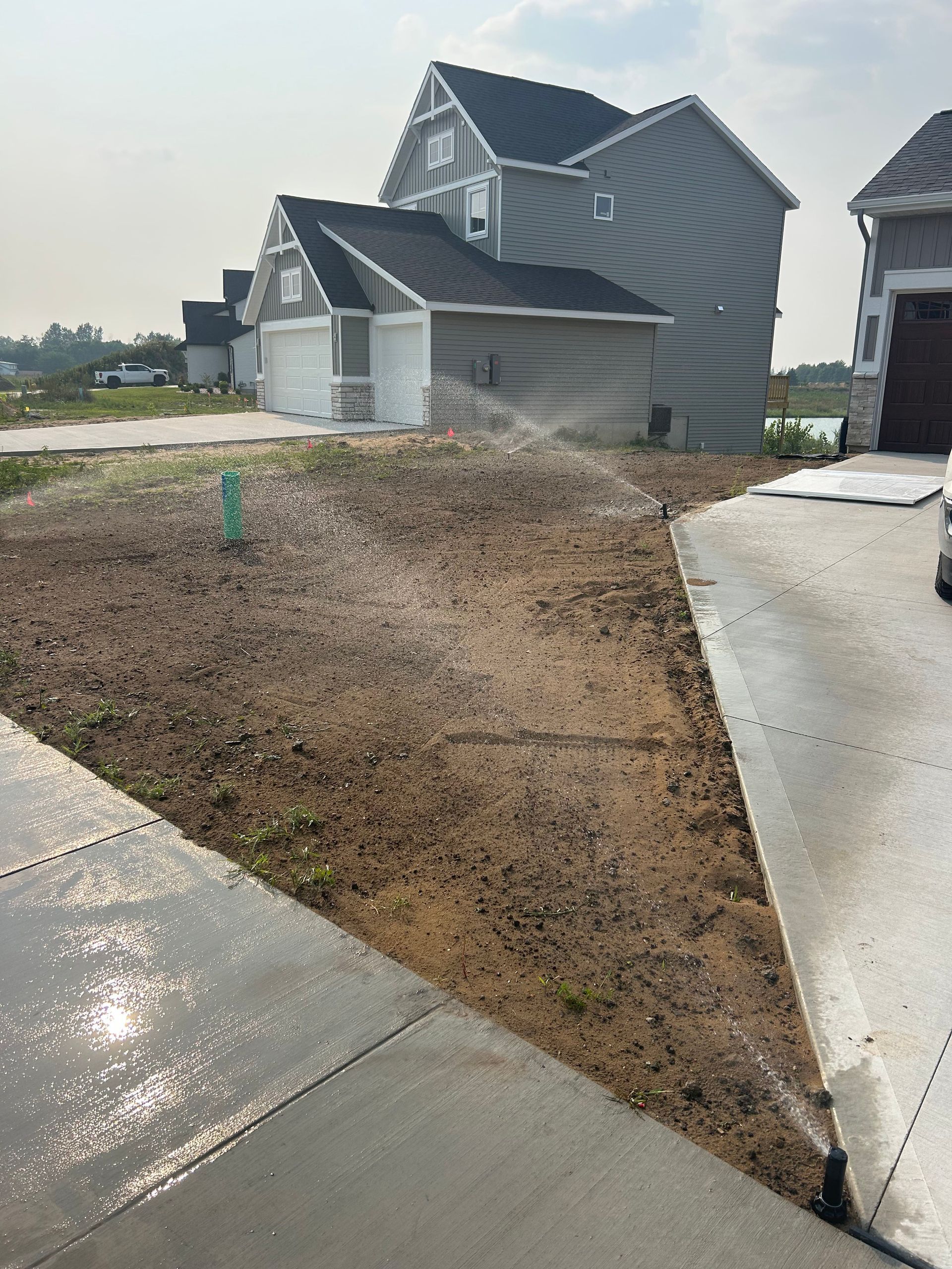 Wet dirt lot beside new gray houses with concrete driveways and a green marker stake