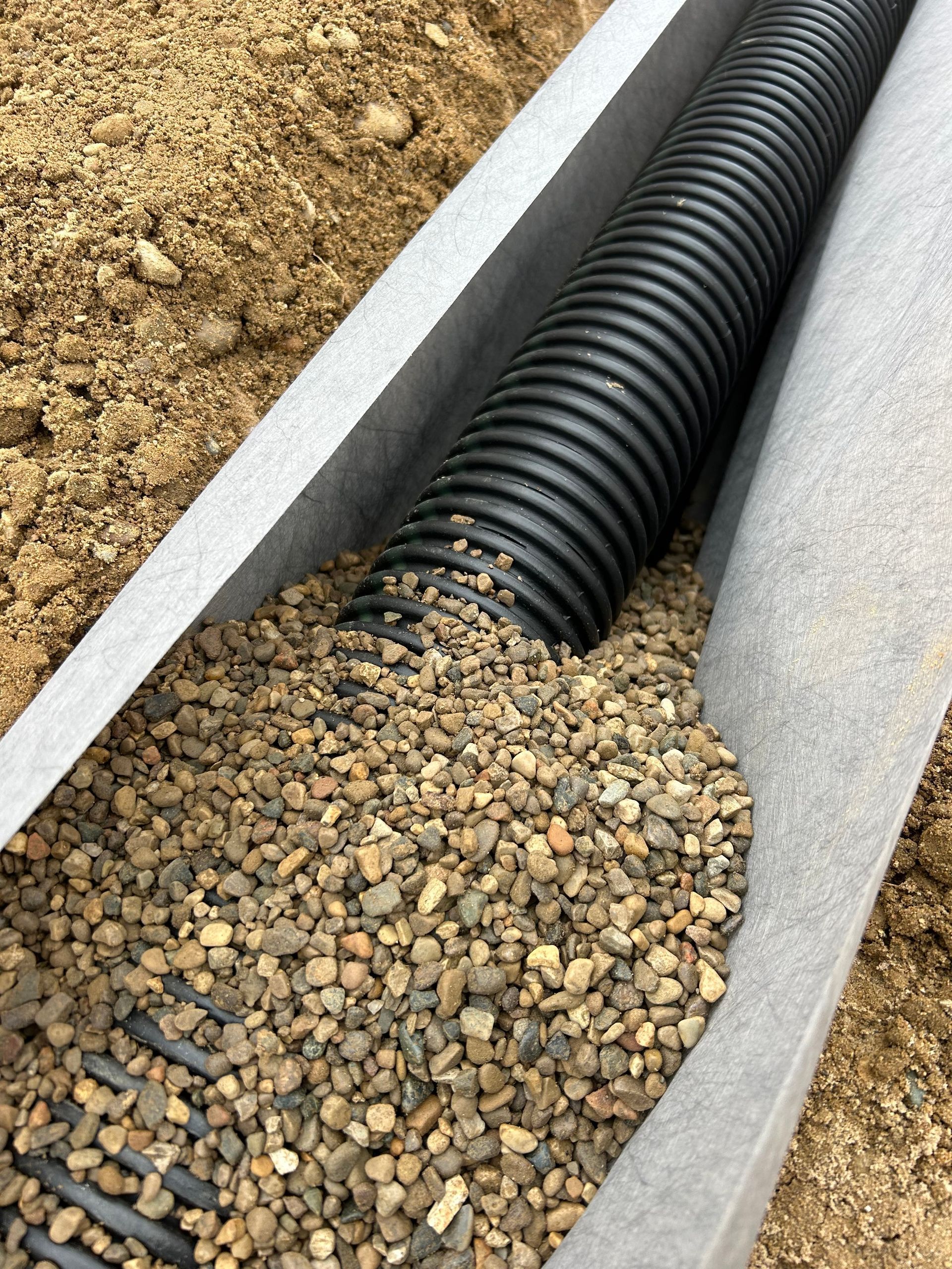 Black drainage pipe in a gravel-filled trench beside soil and concrete edges