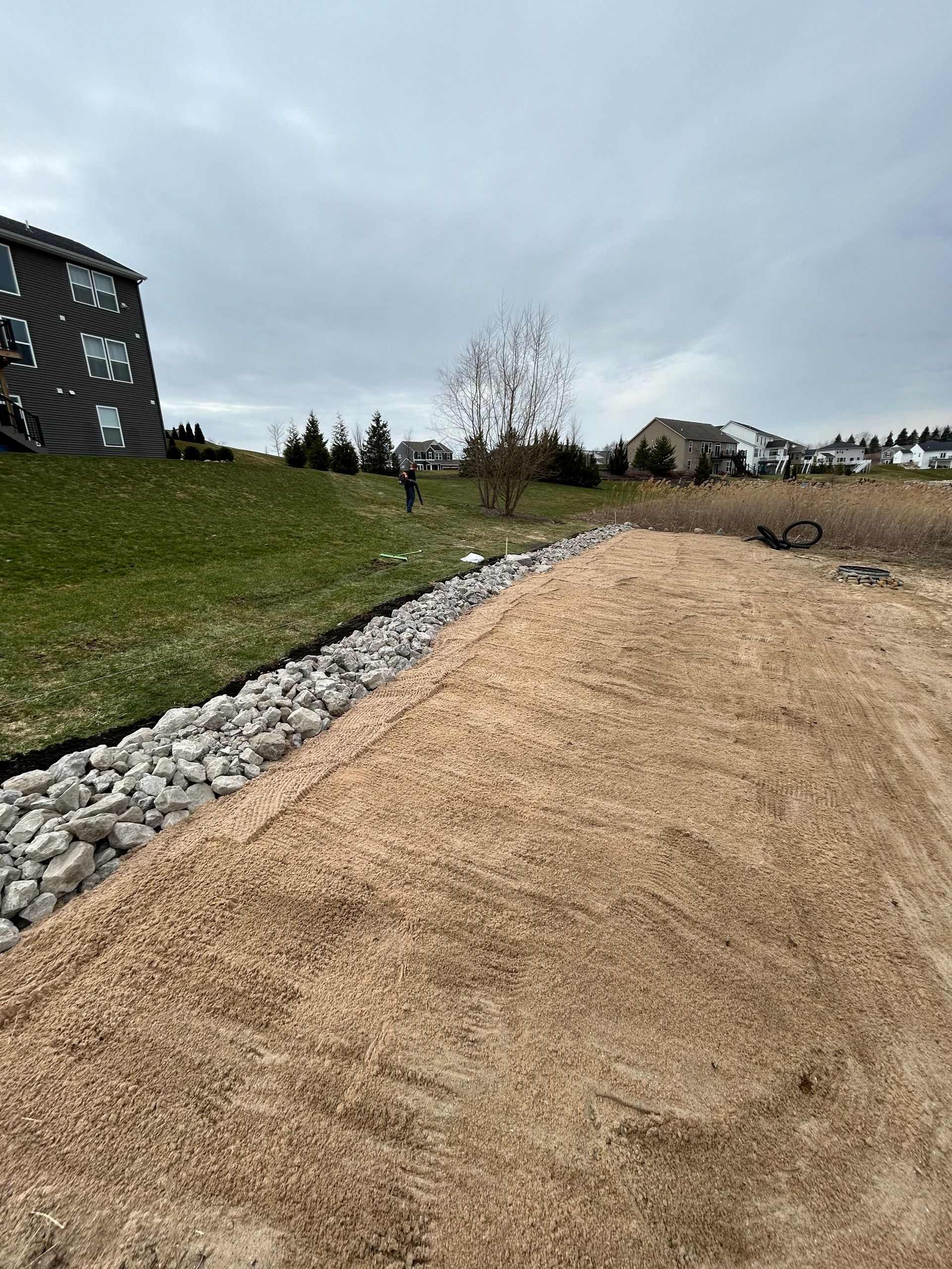 Gravel path beside a rock-lined slope and grassy hill under a cloudy sky, with a building at left