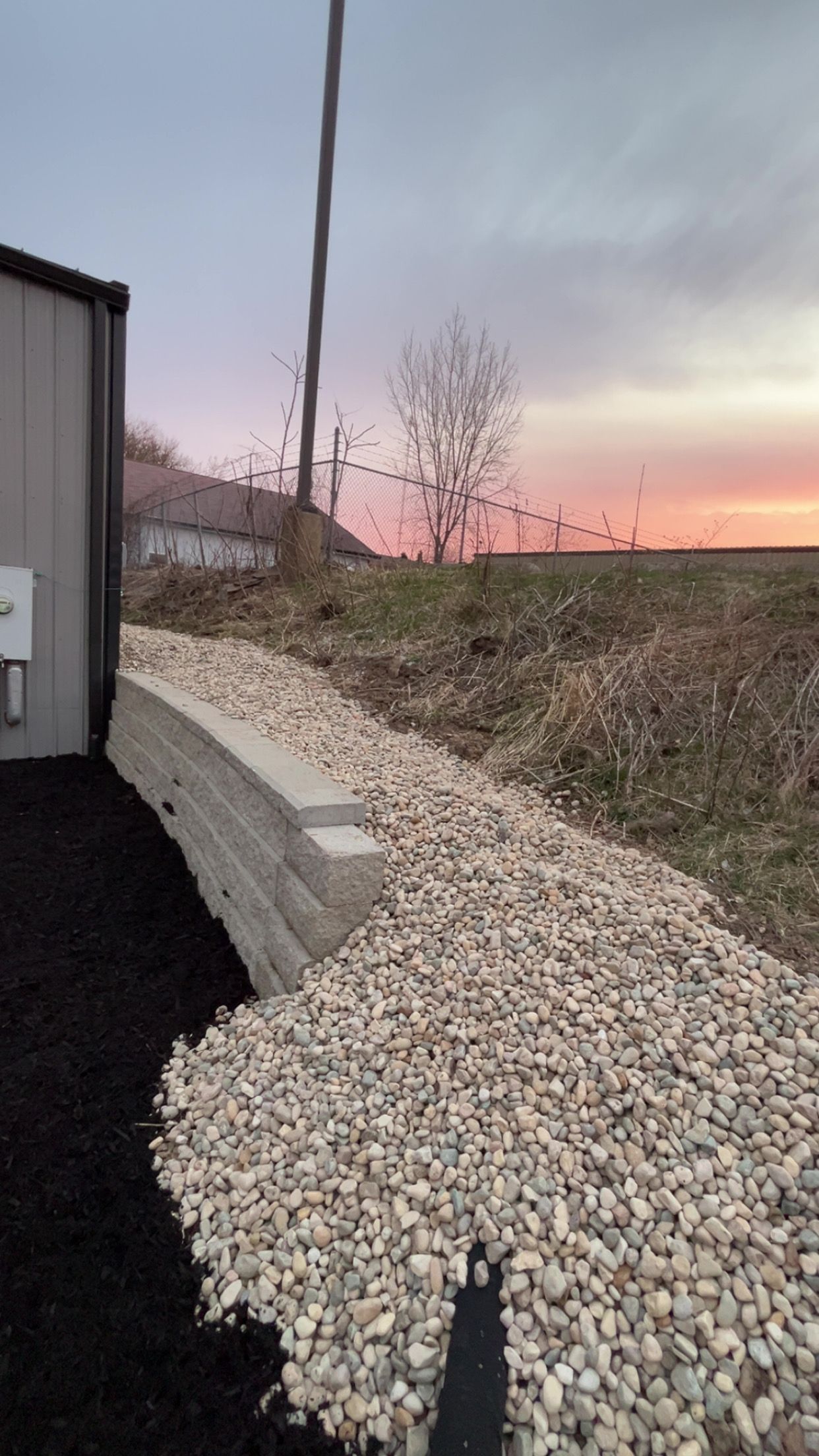 Gravel embankment beside a building and road at dusk, with a utility pole and pink sunset sky.