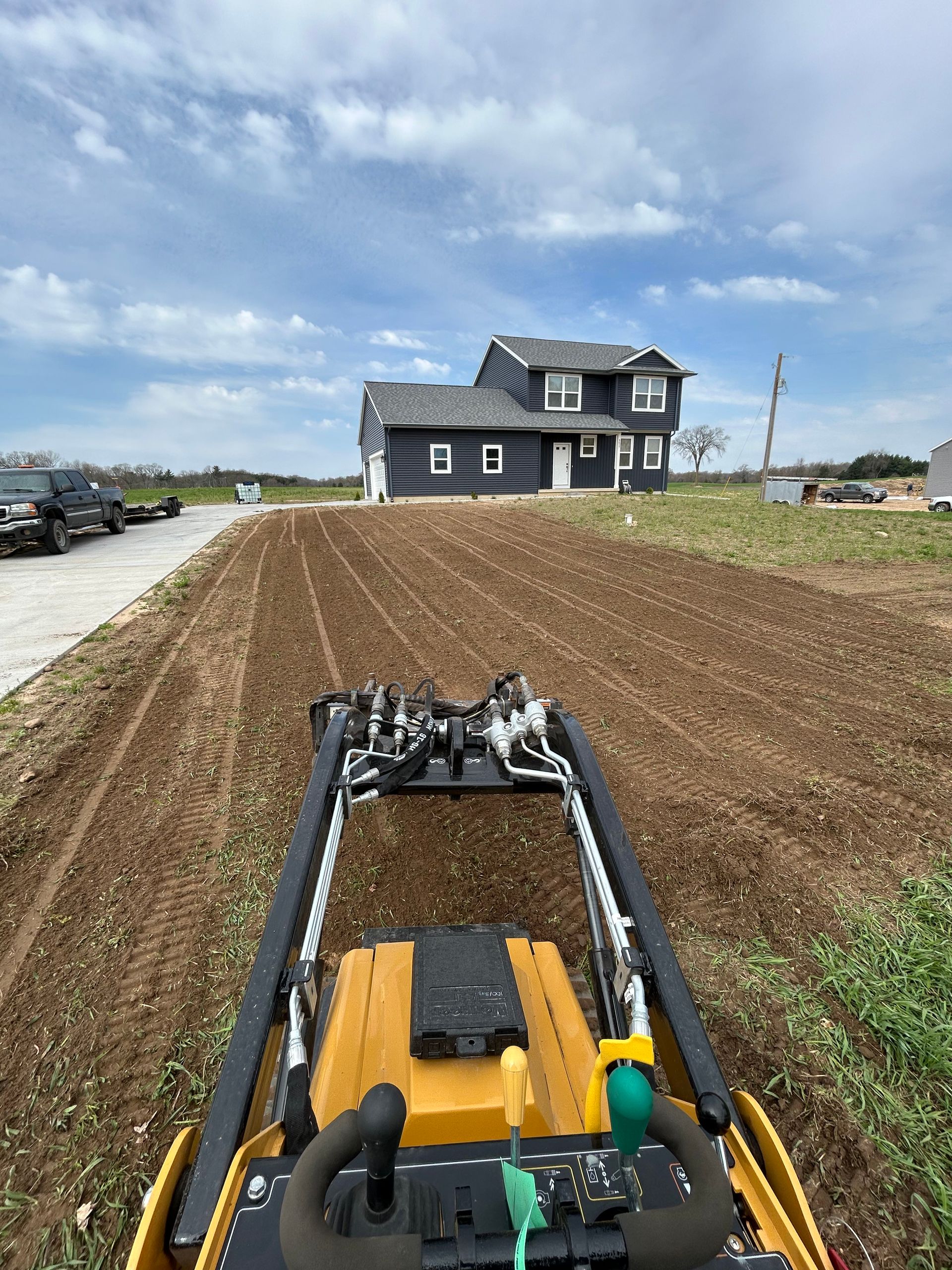 Excavator view of a house under construction on a muddy lot beside a paved road under a cloudy sky