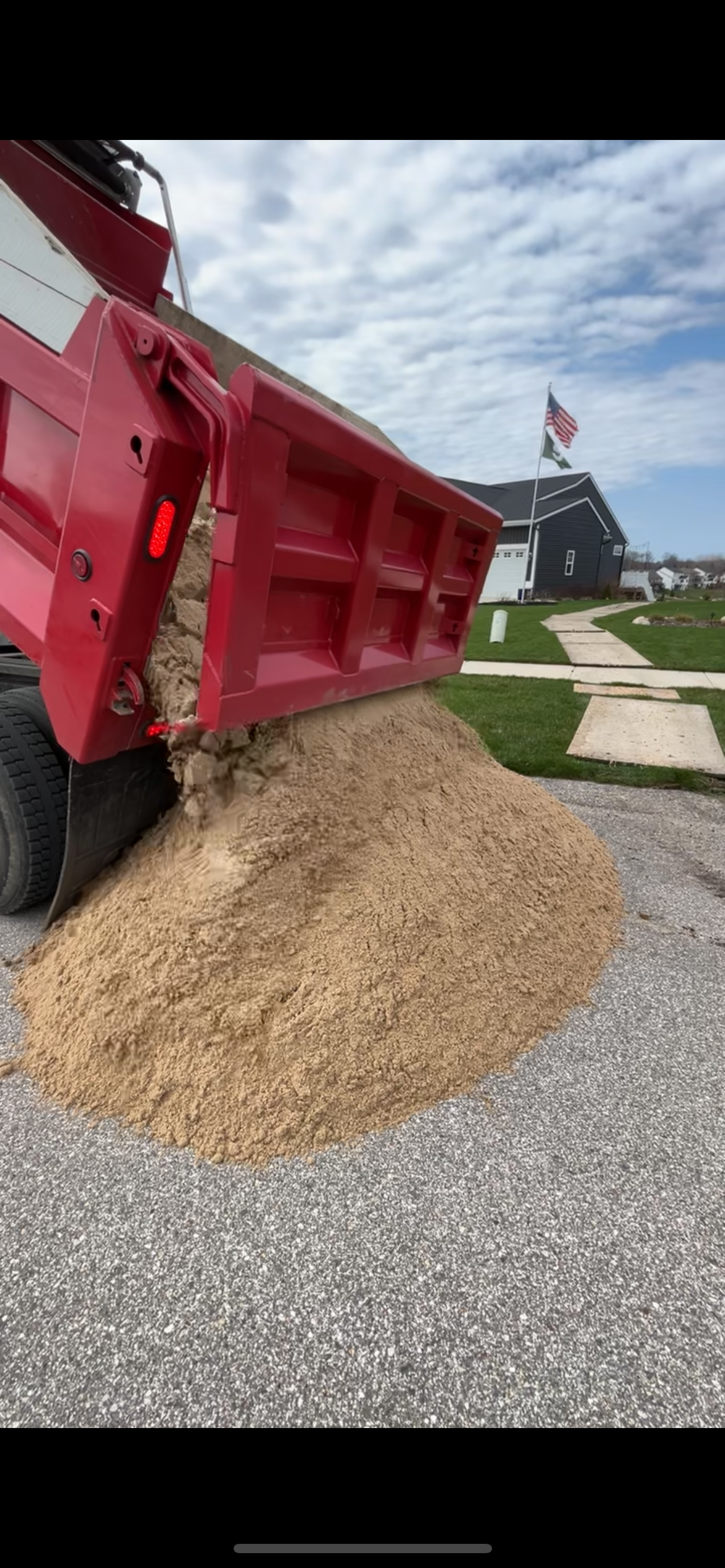 Red dump truck unloading a pile of gravel onto a driveway under a cloudy sky