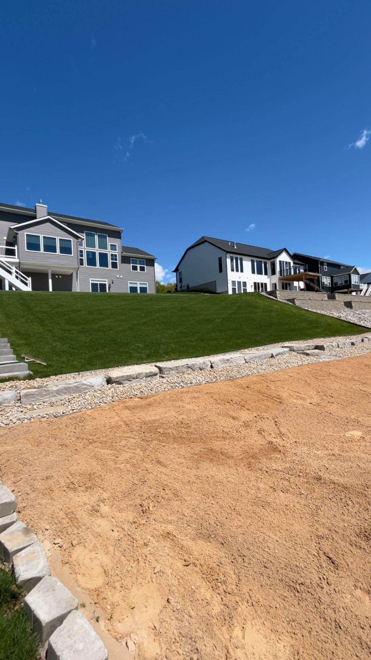 New homes on a grassy slope above a sandy construction lot under a clear blue sky