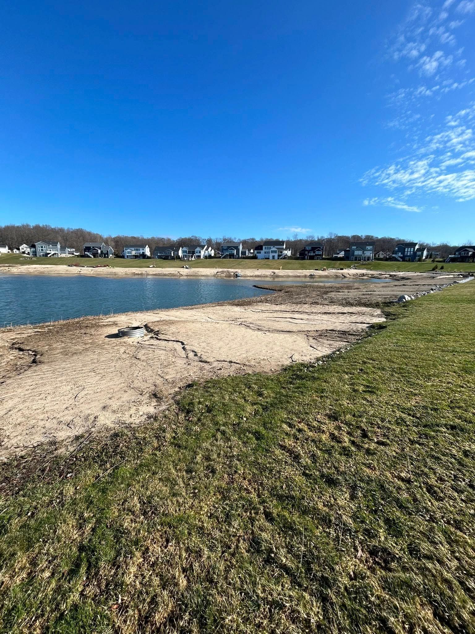 Rocky beach beside a calm lake under a bright blue sky, with houses across the water.