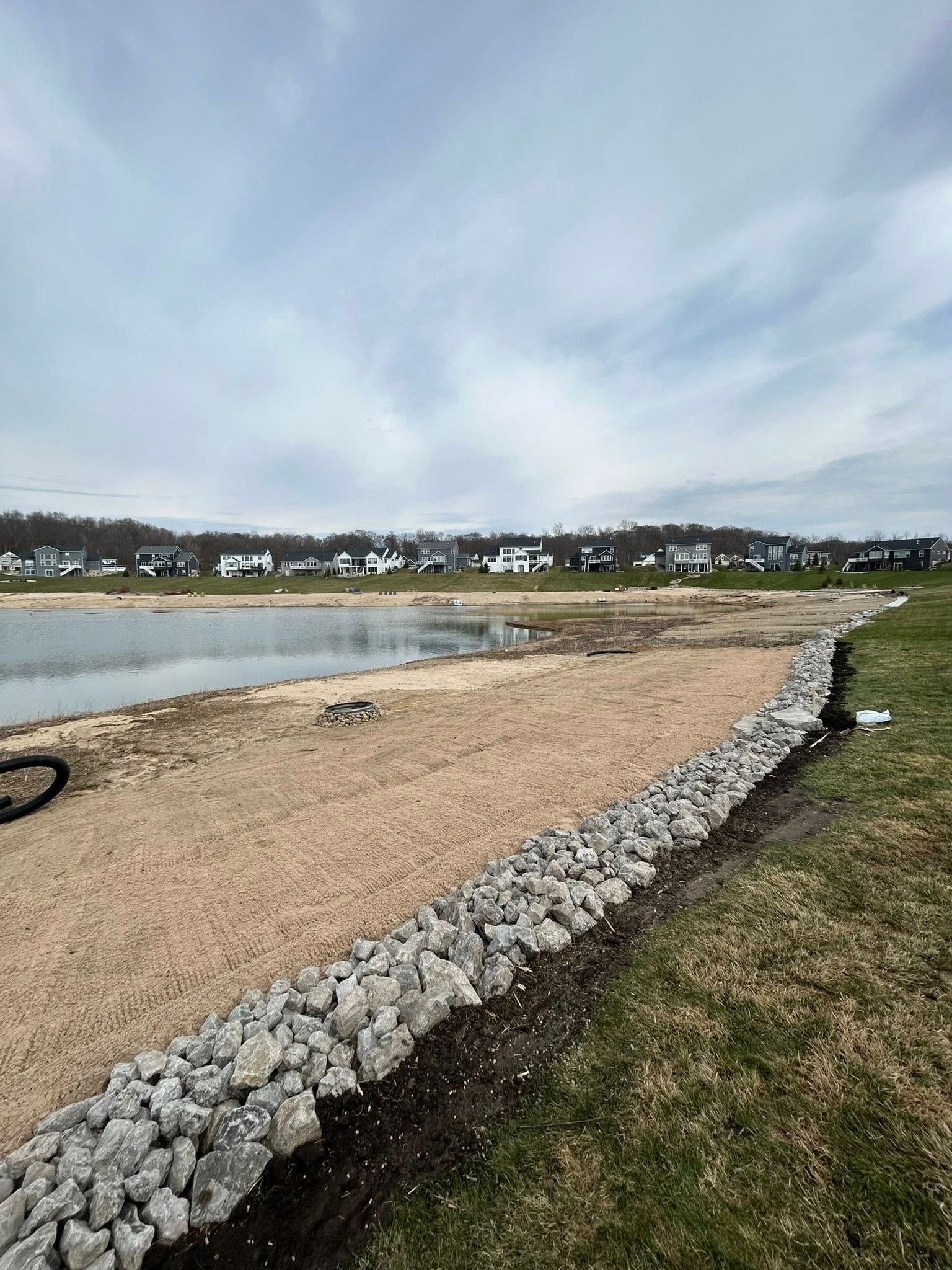 Rock-lined sandy shoreline beside calm water, with houses across the bay under a cloudy sky.