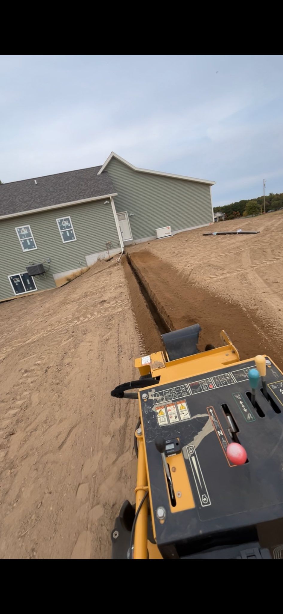 Construction trench beside a house, with a yellow machine and black tool panel in the foreground.